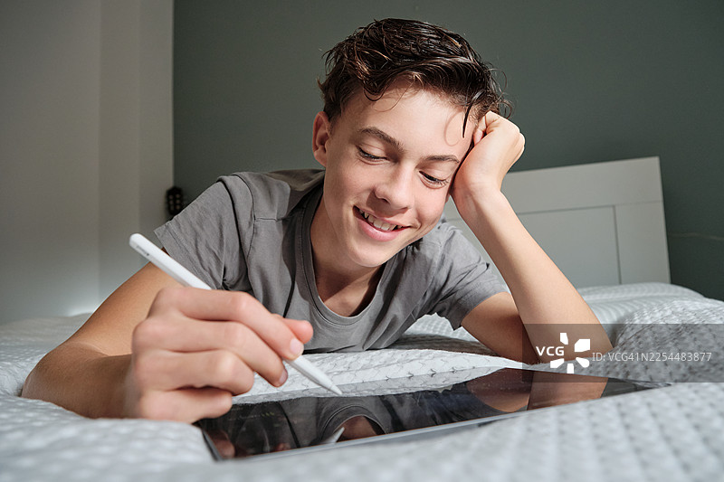 年轻的成年男孩在床上微笑着在数位板上画画图片素材