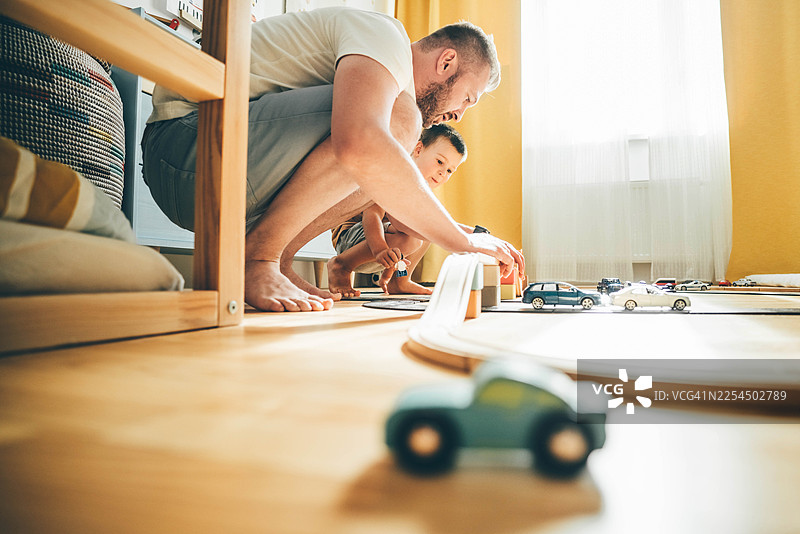 阳光明媚的房间里，父亲和年幼的儿子在地板上玩玩具车。图片素材