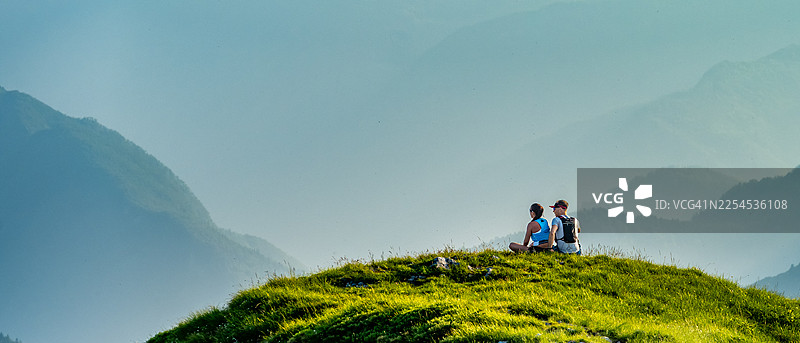 两个人坐在一座绿色的山丘上，眺望着云雾缭绕的山脉图片素材