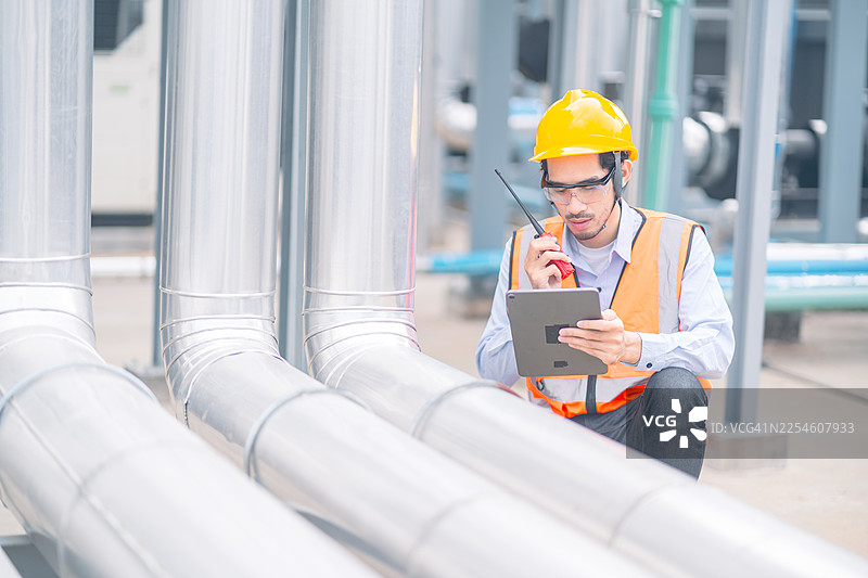 身着安全装备的工程师使用平板电脑和无线电通讯检查工业管道，确保最佳制造效率。图片素材