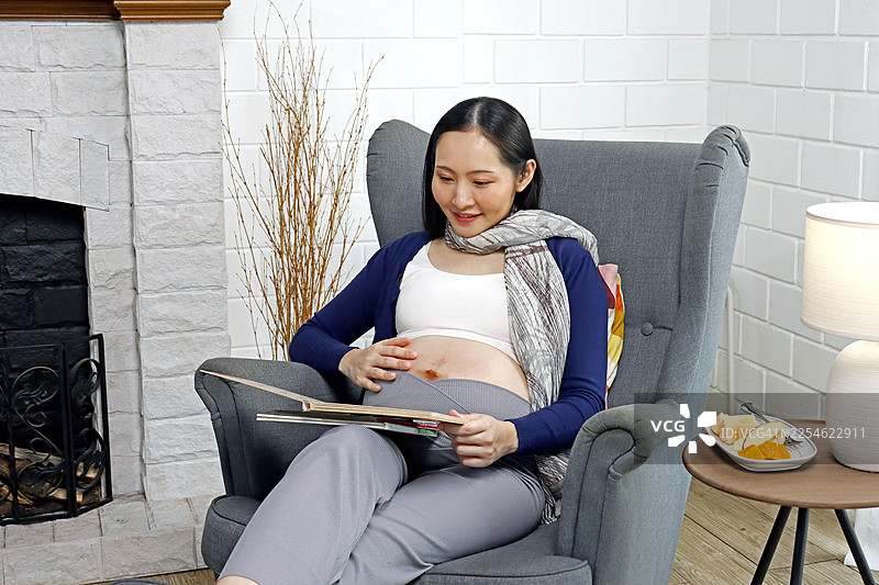 一位快乐的亚洲孕妇在家中舒适的灰色扶手椅上放松，她一边抚摸着肚子一边阅读书籍。图片素材