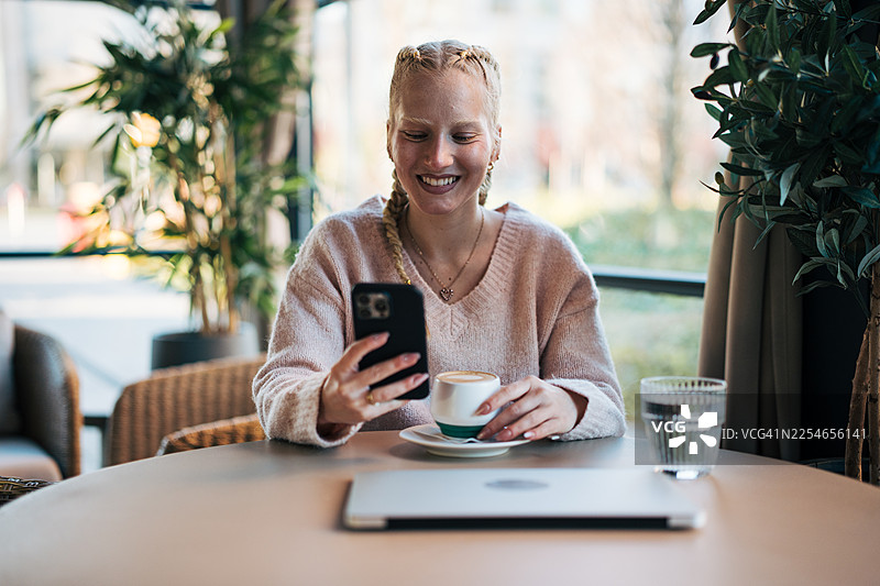 年轻女子在咖啡馆里一边喝咖啡一边微笑着使用手机图片素材