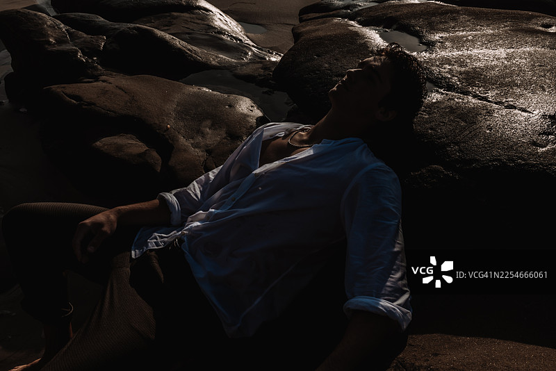 浑身湿透的年轻男子坐在海边的岩石上，海浪拍打着海岸，他正在放松图片素材