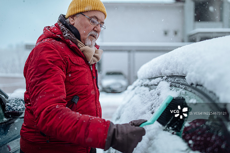 一位穿着红色夹克、戴着黄色帽子的老人在用刷子清理汽车挡风玻璃上的雪图片素材
