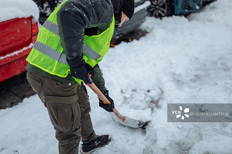 一名穿着高能见度背心的男子在冬季条件下铲除汽车周围的积雪图片素材