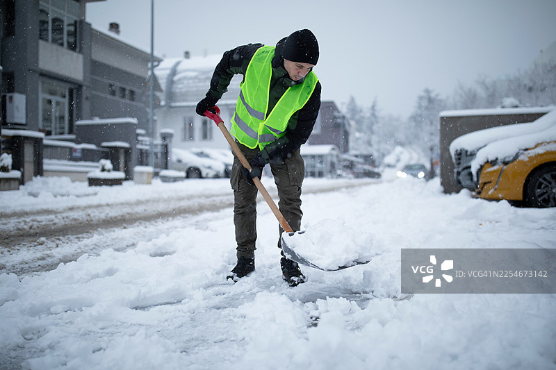 一名身穿高可见度背心的男子在冬季风暴中铲除街道上的积雪图片素材