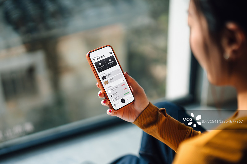 从肩后视角看，年轻的亚洲女性在家中窗边，使用智能手机上的网上银行应用程序管理个人银行和财务。财务规划。投资。财富管理、银行和金融概念。科技赋能智慧银行。图片素材