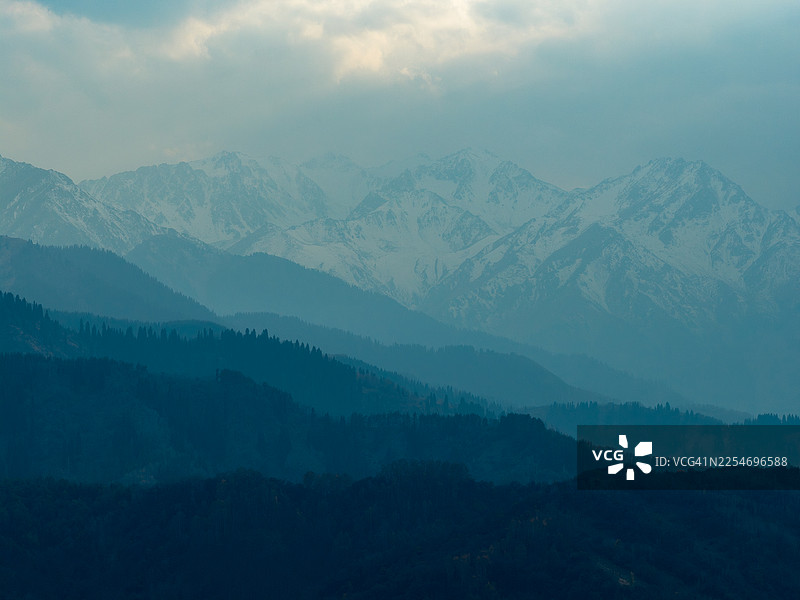 白雪皑皑的壮丽山脉和森林覆盖的山丘在多云的天空下构成了戏剧性而宁静的风景，哈萨克斯坦阿拉木图图片素材