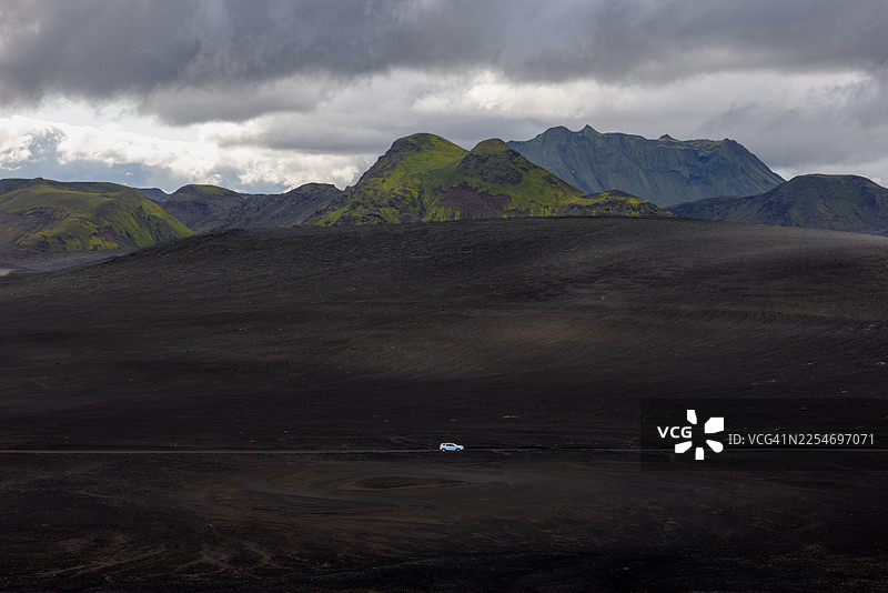 一辆孤零零的车行驶在广阔、黑暗的火山地貌中，苔藓覆盖的山峦在戏剧性的多云天空下若隐若现，地狱，冰岛，兰德曼纳劳卡图片素材