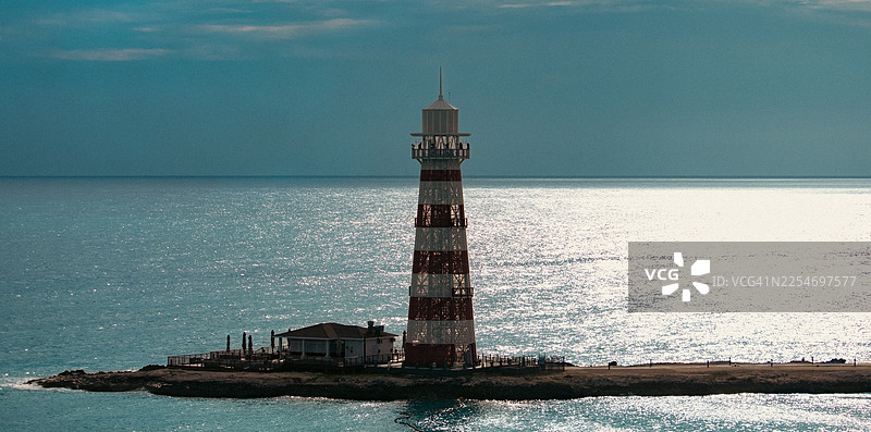 一座红白条纹的灯塔矗立在小岛上，在多云的天空下，它引领着船只驶过波光粼粼的蓝色大海，巴哈马，海洋岛图片素材