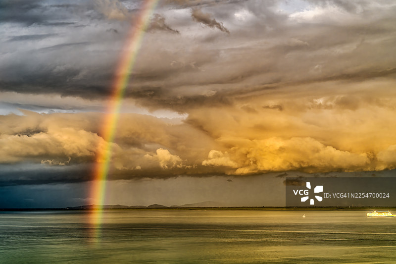 马来西亚丹绒道光，丹绒武雅路，一抹绚丽的彩虹横跨宁静的海面，背景是戏剧性的天空，点缀着金色云彩和远方的陆地图片素材