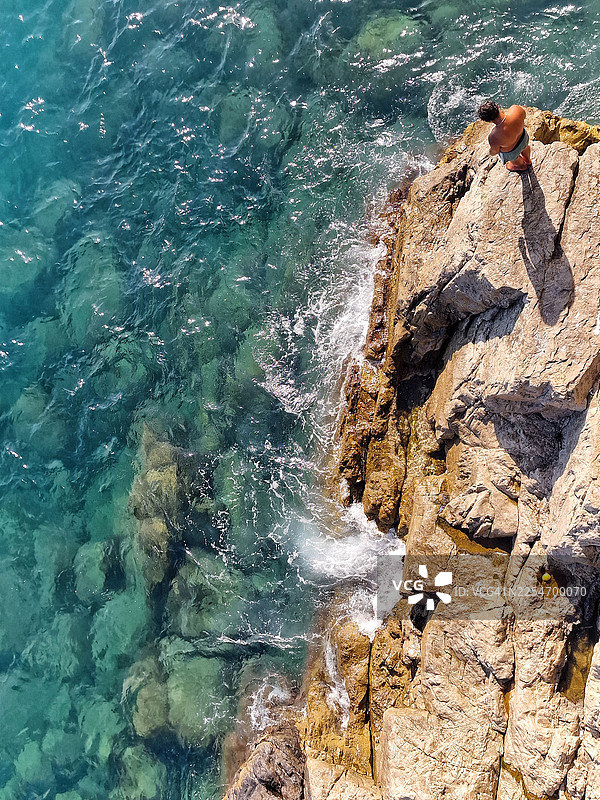 阳光明媚的一天，意大利清澈碧绿的海边，一个人站在崎岖的岩石上，俯瞰视角图片素材