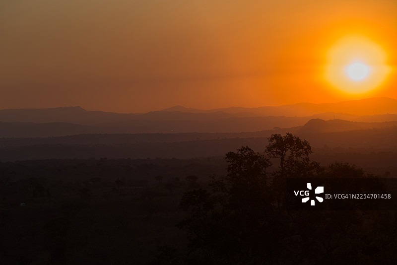 南非姆博贝拉市克鲁格国家公园，山地景观上戏剧性的日落，前景中树木剪影，天空洒满温暖的橙色光辉图片素材