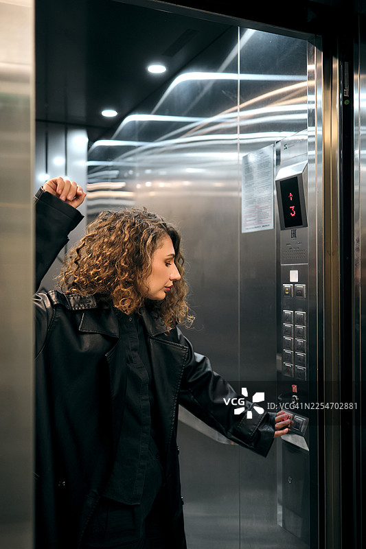 卷发女子身穿黑色外套，在现代电梯里按按钮图片素材