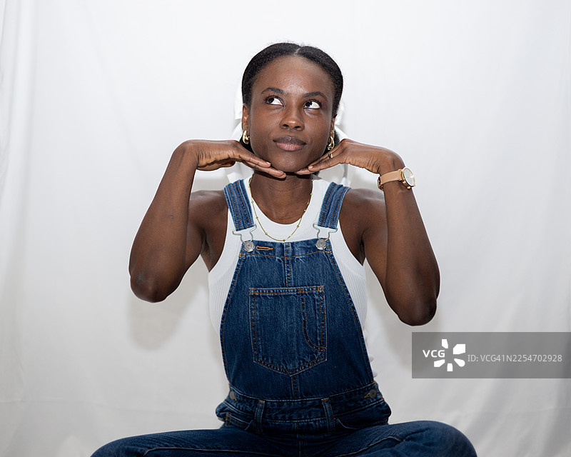 穿着牛仔背带裤的年轻女子，在白色背景前，抬头看向一侧，神情若有所思图片素材