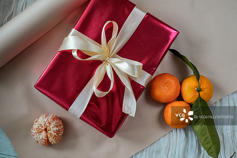 俯视角度拍摄的节日红色礼品盒，带有丝带，周围环绕着新鲜的橘子，背景中性，非常适合节日问候图片素材
