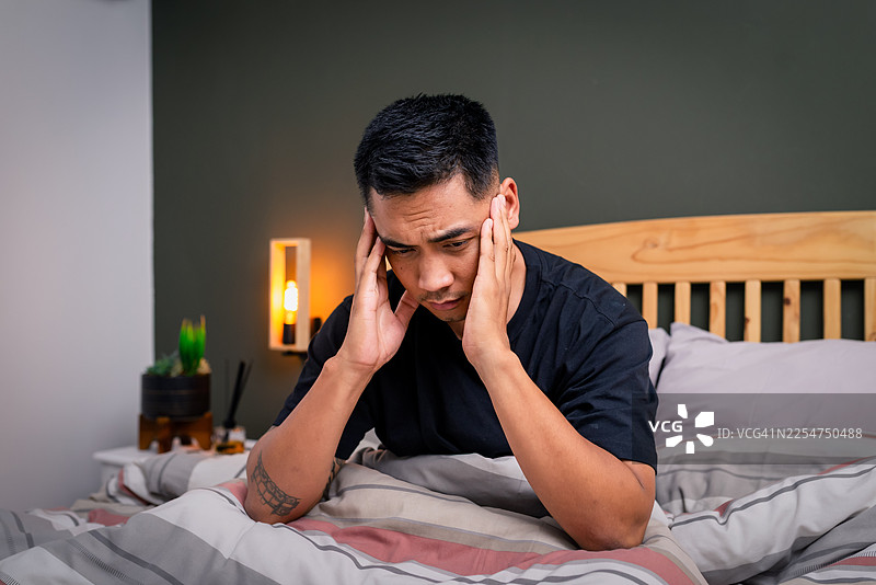 床上男子双手抱头，因压力和疲惫而痛苦，身处舒适的卧室图片素材