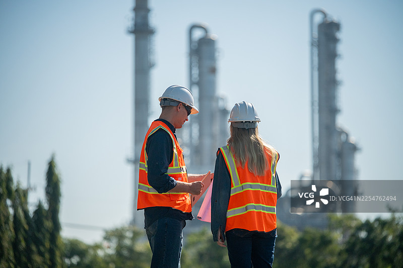 工业工程师戴着安全帽和背心，在平板电脑上检查技术计划和数据，背景是高耸的炼油塔，这是工业安全和碳信用环境运营的概念。图片素材