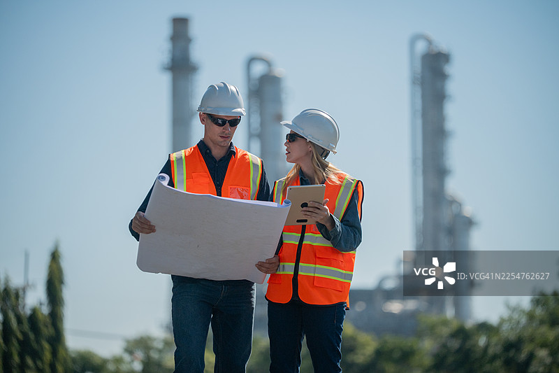 工业工程师佩戴安全帽和背心，在平板电脑上检查技术计划和数据，背景是高耸的炼油塔，这是工业安全和碳信用环境运营的概念。图片素材