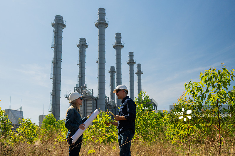身穿安全帽和马甲的工业工程师正在检查平板电脑上的技术计划和数据，背景是高耸的炼油塔，这是工业安全和碳信用环境运营的概念。图片素材