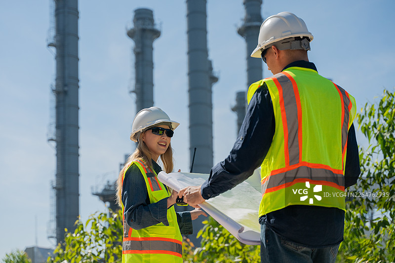 工业工程师佩戴安全头盔和背心，在平板电脑上检查技术计划和数据，背景是高耸的炼油厂塔楼，这是工业安全和碳信用环境运营的概念。图片素材