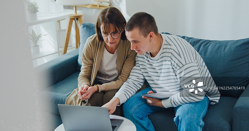 年轻的高加索男子在家中客厅的沙发上教年长的母亲如何使用笔记本电脑。家庭生活方式概念。图片素材
