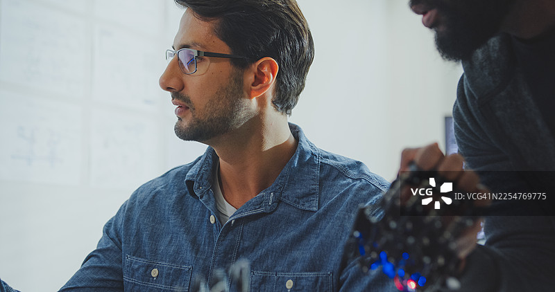 黑人和西班牙裔男性工程师程序员在科技办公室近距离合作，共同研发机器人手部原型，使用计算机控制机器人手臂电路。涉及软件开发、人工智能。图片素材