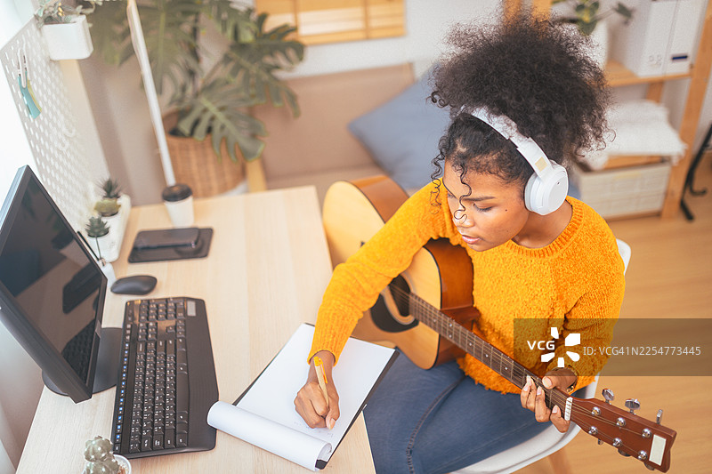 年轻女子在家中用吉他和耳机创作音乐，在舒适的工作空间中激发创造力和提高效率。图片素材
