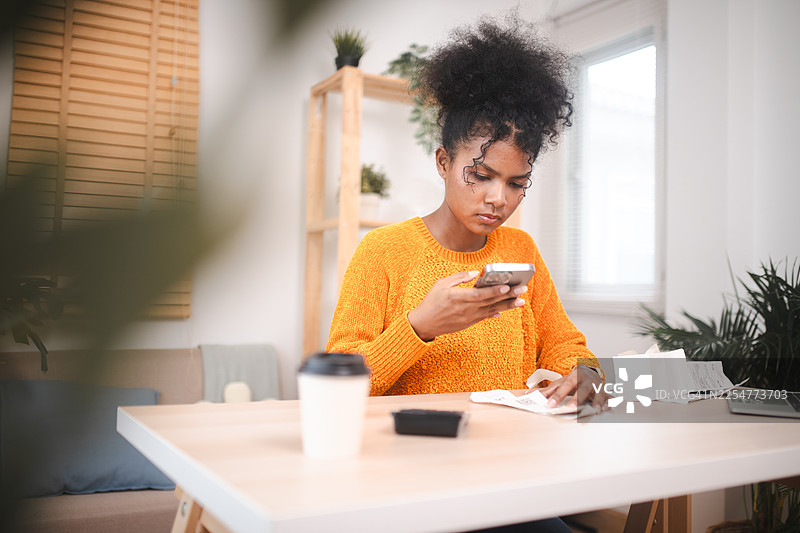 年轻女子在家中的办公室使用智能手机和计算器查看支出，利用科技工具加强财务管理。图片素材