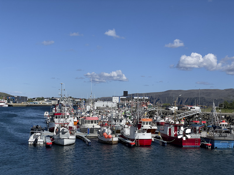 背景为蓝天和群山，阳光明媚的白天，挪威芬马克郡巴特斯峡湾港口停泊着几艘渔船图片素材