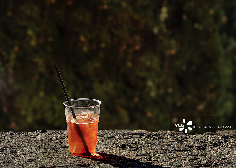 一个装有吸管的塑料杯，里面装着半杯 Spritz 鸡尾酒，放在石头壁架上，沐浴在阳光下。图片素材