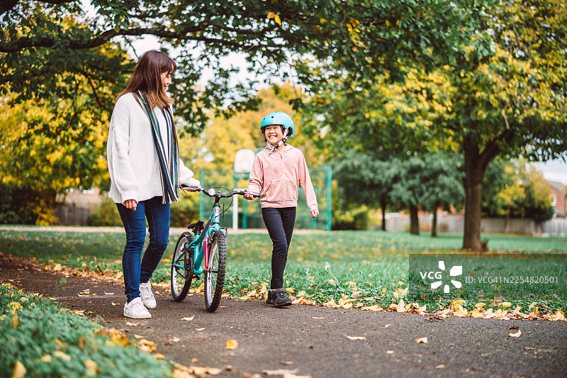 秋日公园里，一位母亲陪伴着女儿学习骑自行车。图片素材