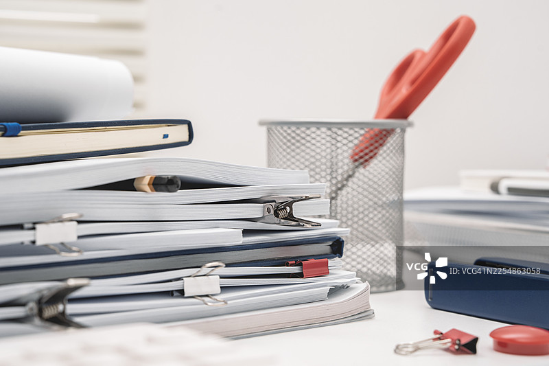 办公桌上的白色文具，堆积如山的文件营造出办公室氛围，还有空白的笔记本。图片素材