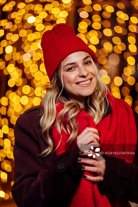 戴着红色绒帽和围巾的微笑女性，背景是温暖灯光的虚化效果图片素材