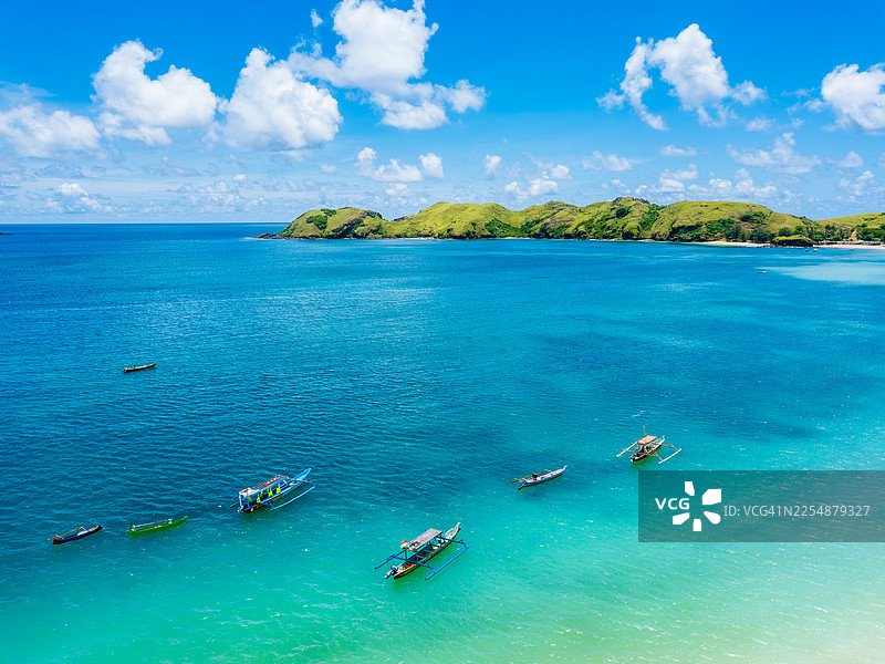 从空中俯瞰，传统的木制支腿船漂浮在绿意盎然的丘陵岛屿附近碧绿的海水中，该岛位于印度尼西亚龙目岛。图片素材
