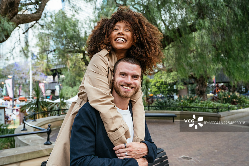 一对快乐的拉丁情侣在城市公园里携手漫步，公园靠近水滨，背景是林立的城市摩天大楼。图片素材