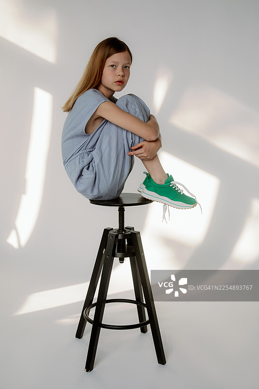 身穿蓝色连体裤和绿色运动鞋的女孩坐在录音室凳子上，柔和光线下平静的肖像照图片素材