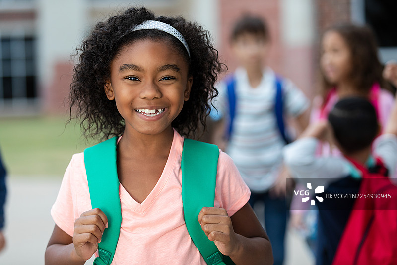 一个背着背包的快乐的年轻黑人女孩对着镜头微笑，背景中的其他学生模糊不清图片素材