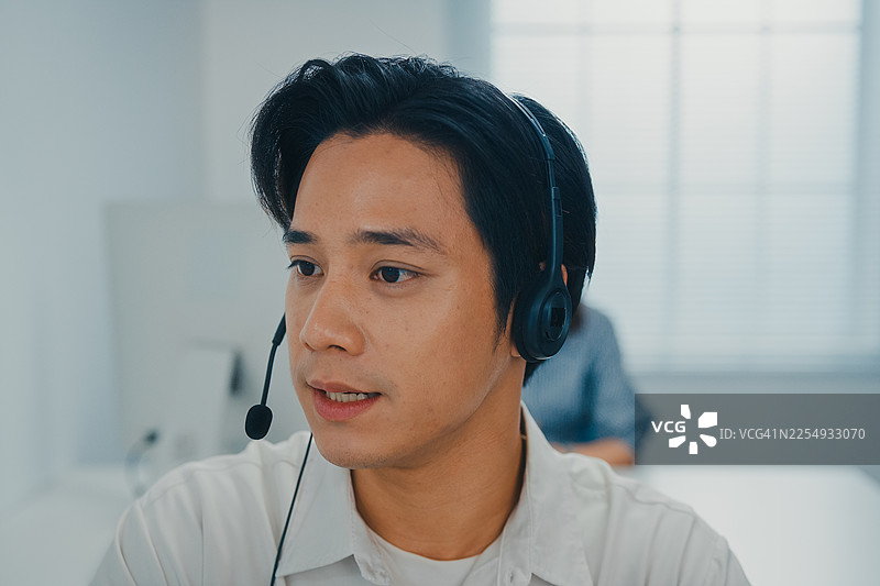 近距离拍摄的年轻亚洲客服代表佩戴耳机，正在协助客户专员在呼叫中心办公室解决客户请求。专业的办公数据中心。服务台操作员。图片素材