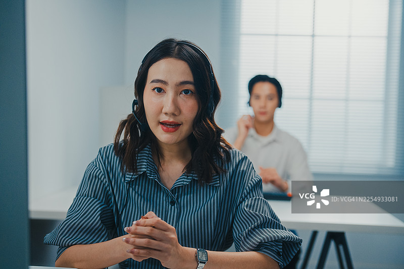 年轻的亚裔客服代表戴着耳机，在呼叫中心办公室协助客户专家解决客户请求。专业数据中心工作。服务台操作员概念。图片素材