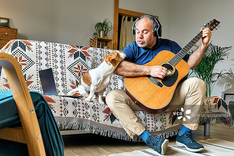 一位留着胡须的中年男子坐在客厅的沙发上，为他的杰克罗素梗犬弹奏木吉他。图片素材
