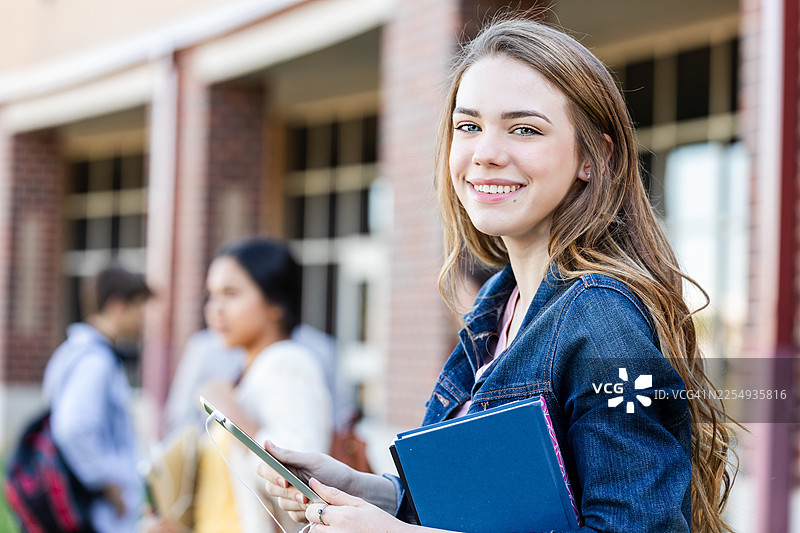 年轻女子在教学楼外微笑着，手中拿着书籍和数码平板电脑图片素材