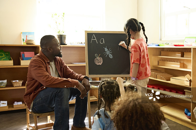 老师看着女孩在黑板上写字母图片素材