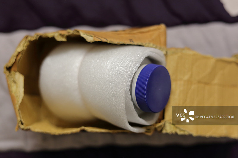 一个带有蓝色瓶盖的瓶子，用蓬松的白色泡沫纸包裹，放在一个已打开的快递纸箱中以确保安全。图片素材
