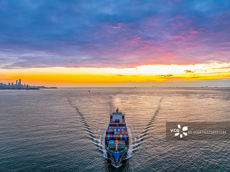 日出时分，无人机航拍视角下的集装箱货轮，在青岛城市天际线前，航行于绚丽的天空和彩色的云彩之下图片素材