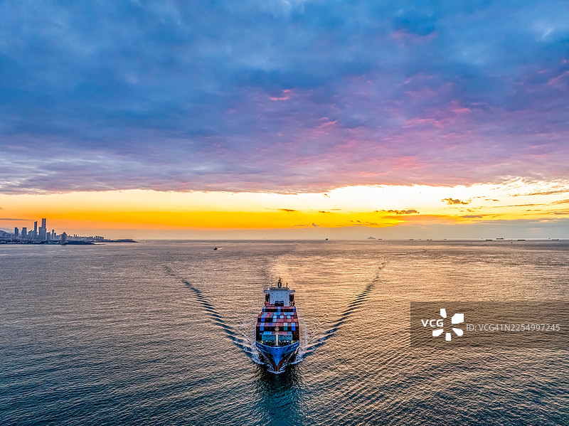 无人机航拍，日出时分，在青岛城市天际线前，一艘集装箱船正航行在美丽的天空和多彩的云彩下图片素材