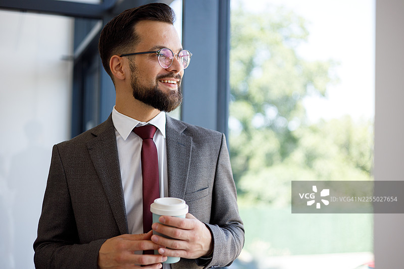 戴眼镜的年轻商人微笑着，手里拿着咖啡杯，透过窗户眺望，思考着成功图片素材