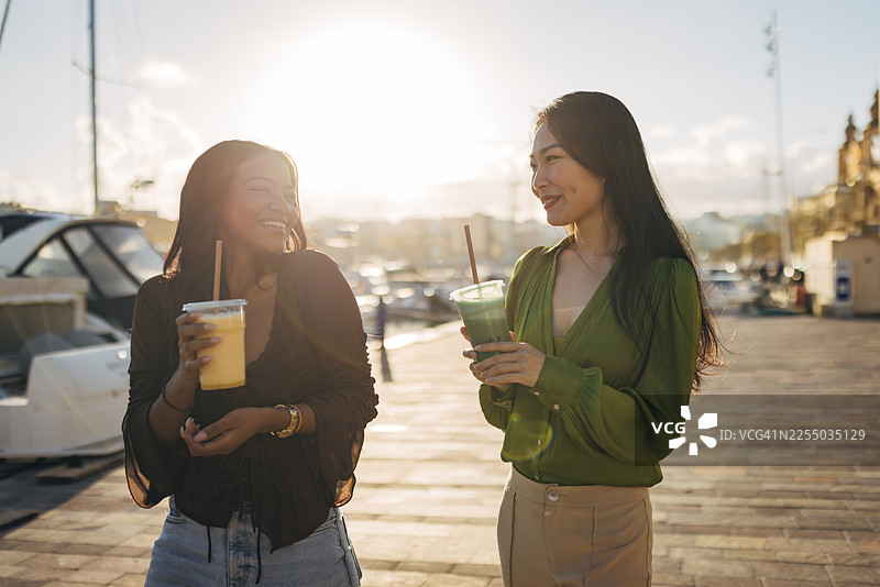 两位开心的女士在海滨散步，享用新鲜水果冰沙。她们一边笑一边聊天，展现了度假期间健康积极的生活方式。图片素材