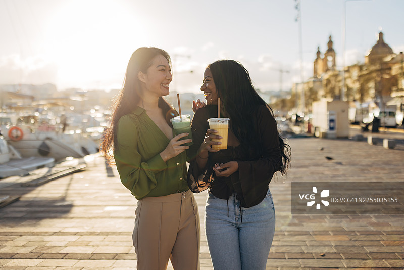 两位不同种族的好朋友在阳光明媚的石板路海滨散步，她们手里拿着冰镇水果冰沙，互相看着对方大笑，在旅途中庆祝友谊。图片素材