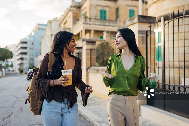 两位不同肤色的女性在风景如画的城市街道上边走边笑谈。她们手中拿着健康的即饮饮品，庆祝着她们的友谊和旅行的乐趣。图片素材